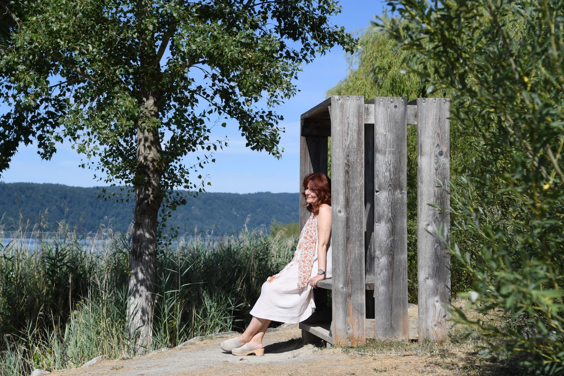 Sabine Jahn sitzt lächelnd in einem Holzpavillon am Seeufer mit Blick auf Schilf und See